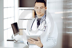 Friendly male doctor using tablet computer in sunny clinic. Medicine and healthcare concept