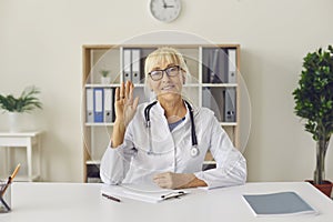 Friendly mature online doctor waving hand at camera sitting at desk in hospital office