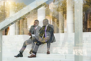 Friendly meeting of business partners outdoors. Two dark-skinned men in suits sit on the steps of a city building with a