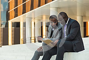 Friendly meeting of business partners outdoors. Two dark-skinned men in suits sit on the steps of a city building with a