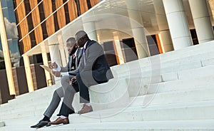 Friendly meeting of business partners outdoors. Two dark-skinned men in suits sit on the steps of a city building with a