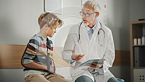 Friendly Middle Aged Family Doctor Talking with a Young Boy with Arm Brace and Showing Test Results