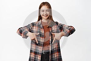 Friendly milennial girl smiling, pointing fingers down, showing announcement, advertisement or logo, indicating banner