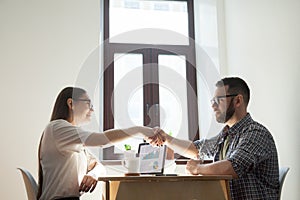 Friendly millennial coworkers handshaking over office desk. Busi