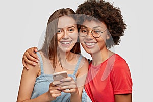 Friendly mixed race women embrace and smile together, watch funny video content on cellular, pose against white