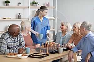 Friendly nurse holding mugs, talking to elderly people, serving tea