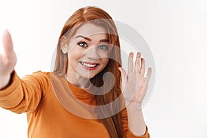 Friendly and outgoing pretty redhead woman in orange sweater, extend arm holding smartphone or camera, taking selfie