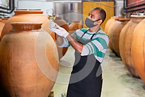 Friendly owner in protective mask of winery standing with wine in wooden barrels cellar