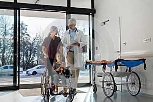 Friendly pediatrician nurse talking, pushing little patient in wheelchair. Cute preschool girl trusting her doctor and