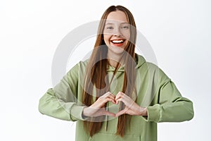 Friendly positive girl with long hair, show heart i love you gesture, smiling and looking happy at camera, standing in