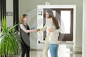 Friendly realtor and young couple shaking hands standing in hall