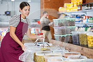 Friendly saleswoman in an apron offers to buy pickled green olives in grocery supermarket