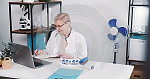 Friendly senior Caucasian doctor woman in lab coat, eyeglasses smiles consulting remote patient using laptop webcam chat