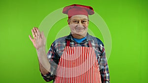 Friendly senior chef in red apron waves palm for greeting or goodbye sign.