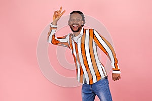 Friendly smiling african man with dreadlocks in trendy striped shirt and denim pants showing victory gesture with fingers, looking