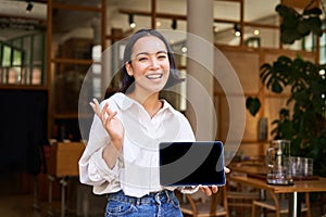 Friendly smiling asian woman, cafe manager, showing tablet screen and looking happy, demonstrating her restaurant profit