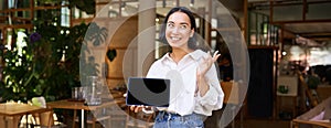 Friendly smiling asian woman, cafe manager, showing tablet screen and looking happy, demonstrating her restaurant profit