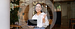 Friendly smiling asian woman, cafe manager, showing tablet screen and looking happy, demonstrating her restaurant profit