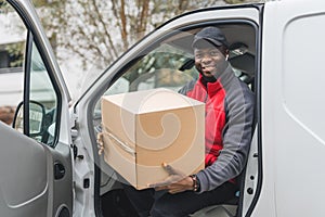 Friendly smiling Black man in his mid 20 sitting on driver's seat with opened door and holding cardboard package