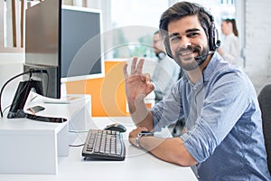 Friendly smiling customer support operator with hands-free headset showing okay gesture in the office.