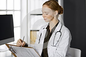Friendly smiling female doctor using clipboard in clinic. Portrait of friendly physician woman at work place. Perfect