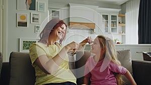 A friendly smiling mother sits on the sofa in the room with her daughter, braids her hair for preparation to the matinee