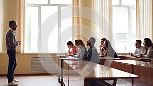 Friendly teacher is talking to students, smart mixed race man is raising hand and speaking sitting at table in lecture