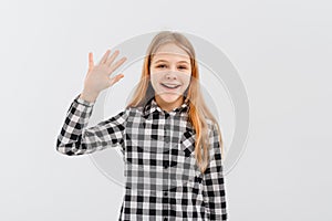 Friendly teen girl says hello, waves hand and smiles happy at you, stands over white background in casual shirt