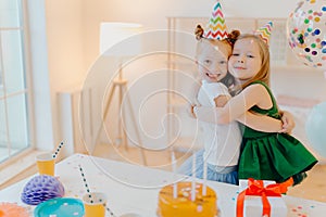 Friendly two girls embrace and have good relationship, stand near festive table with cake, celebrate birthday together, stand in