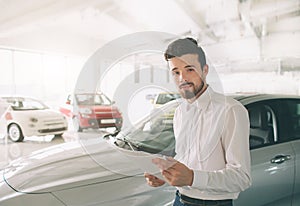 Friendly vehicle salesman presenting new cars at showroom. Photo of young male consultant showing new car in auto show