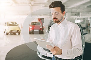 Friendly vehicle salesman presenting new cars at showroom. Photo of young male consultant showing new car in auto show