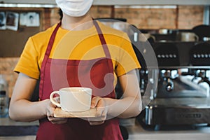 Friendly waitress woman wearing protection face mask waiting for serving hot coffee cup to customer in cafe coffee shop, cafe rest