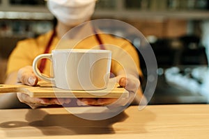 Friendly waitress woman wearing protection face mask waiting for serving hot coffee cup to customer in cafe coffee shop, cafe rest