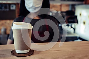 Friendly woman barista or waitress wearing protection face mask waiting for serving hot coffee cup to customer in cafe coffee shop