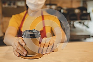 Friendly woman barista or waitress wearing protection face mask waiting for serving hot coffee cup to customer in cafe coffee shop