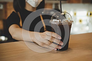 Friendly woman waitress wearing protection face mask waiting for serving ice black coffee to customer in cafe coffee shop