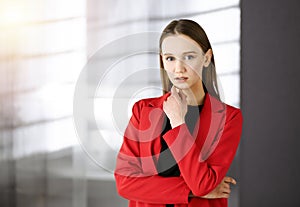 Friendly young business woman or female student dressed in red coat is standing straight and looking at camera in a