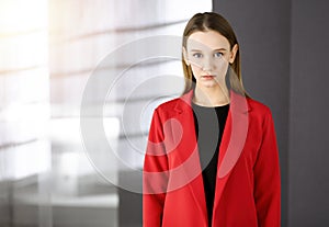 Friendly young business woman or female student dressed in red coat is standing straight and looking at camera in a