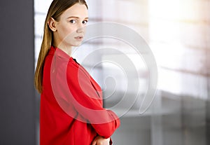 Friendly young business woman or female student dressed in red coat is standing straight and looking at camera in a
