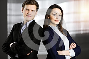 Friendly young colleagues are standing as a team with crossed arms in sunny office. Successful business people at work