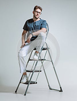 Friendly young male workman repairman in uniform, doctor sitting on stepladder. Confident guy on ladder, studio portrait