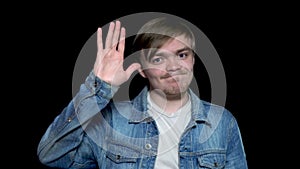 Friendly young man in jean jacket waving his hand to say goodbye, isolated on black background. Polite young man saying