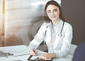 Friendly young woman-doctor is listening to her patient, while sitting together in sunny clinic. Medicine and healthcare