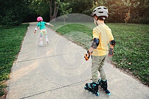 Friends boy and girl in helmets riding on roller skates in park on summer day. Sister encourage stimulate brother to ride on