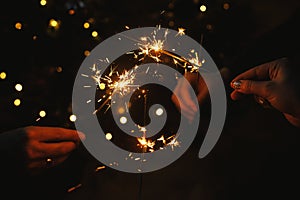 Friends celebrating with burning sparklers in hands against christmas lights in dark room. Happy New Year! Atmospheric holiday.