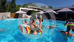 Friends chilling in private villa swimming pool, lie in the sun on inflatable flamingo, swan, floaties. Young people