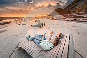 Friends communicate by smartphone and relax lying on a bench in the city Park on the river embankment.