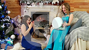 Friends exchange Christmas presents New Year`s Eve, joyful beautiful female give a gift, girls sit by the fireplace