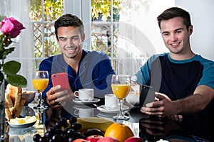 Friends or Gay couple sitting at breakfast table in front of patio doors looking at mobile devices and smiling