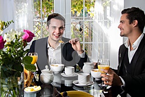 Friends or Gay couple sitting at table in front of patio doors eating breakfast with one man looking at camera